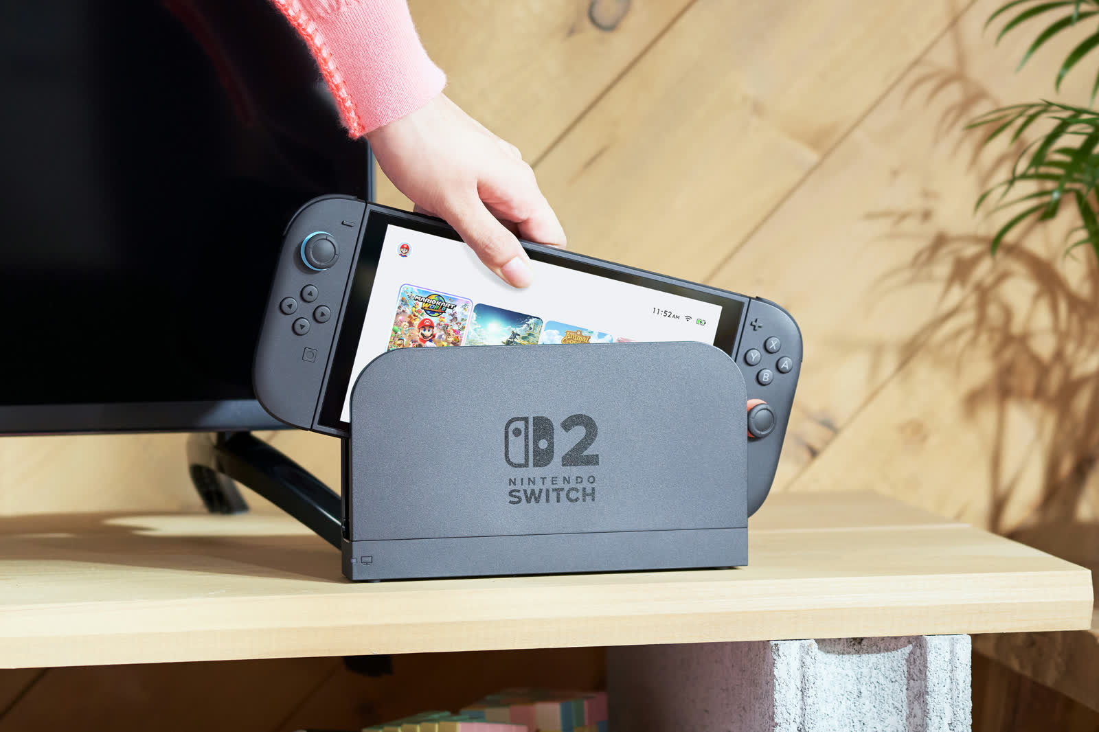 Oficjalne zdjęcie Nintendo — konsola Nintendo Switch 2 na podstawce na biurku, obok dłonie trzymające kontroler Joy-Con 2